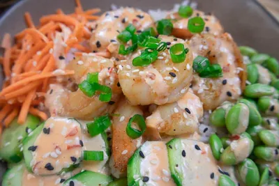 Shrimp poke bowl with carrots, cucumber, edamame, sesame seeds, and spicy mayo, garnished with green onions.
