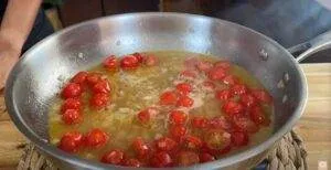 Cherry tomatoes simmering in a skillet with garlic and olive oil on a wooden surface.