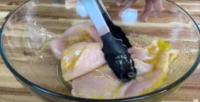 Tongs mixing raw chicken breast in marinade in a glass bowl on a wooden table.