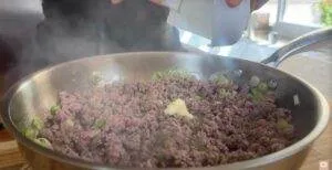 Cooking ground beef with scallions and spices in a skillet. Hands adding seasoning for a flavorful dish.
