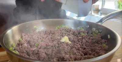 Cooking ground beef with scallions and spices in a skillet. Hands adding seasoning for a flavorful dish.