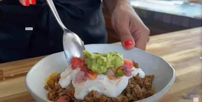 taco potato bowl with guacamole, sour cream, and salsa being prepared with a spoon on a wooden table.