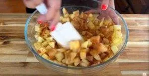 Mixing diced apples with cinnamon in a glass bowl for pie preparation.