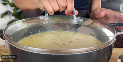 Person lifting lid off a pot of creamy soup in a kitchen setting.