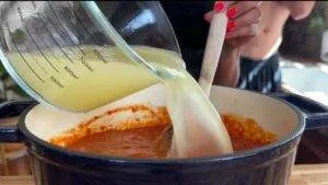 Pouring chicken broth into a simmering pot of tomato sauce, stirred by a wooden spoon.