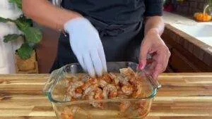 Person marinating shrimp with spices in a glass dish, wearing gloves, on a wooden kitchen counter.