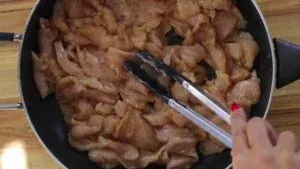 Hand stirring sliced raw chicken in a pan with tongs on a wooden surface.