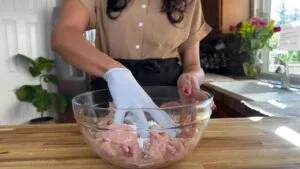 Person marinating chicken in a glass bowl with gloves in a kitchen setting.
