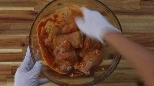 Gloved hands mixing marinated chicken pieces in a glass bowl on a wooden surface.