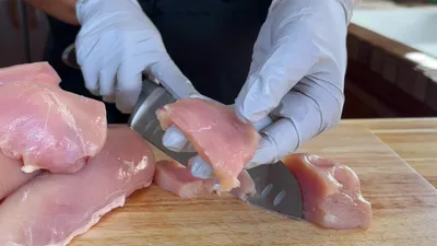 Gloved hands slicing raw chicken on a wooden cutting board for meal preparation.