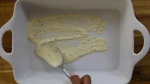 Spreading creamy sauce in a white baking dish with a spoon for a delicious recipe.