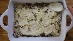 Layered casserole with cheese and sauce being prepared in a white baking dish on a wooden surface.