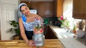 Woman using a food processor in a cozy kitchen with wooden cabinets and floral decor.