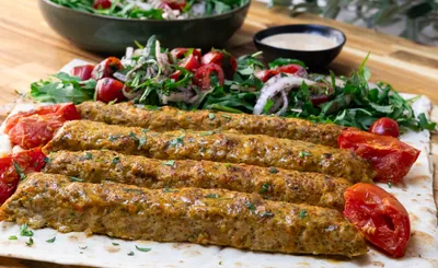 Grilled kebabs with herbs on flatbread, served with fresh tomato salad and a side of sauce.