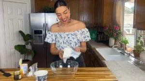 Woman preparing ingredients in a bright kitchen, holding cheese by a bowl on a wooden counter with flowers nearby.