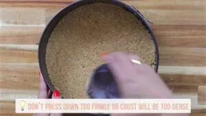 Person pressing graham cracker crust into a round pan with a glass on a wooden surface.