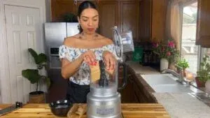 Woman using a food processor in a kitchen with plants and flowers on the counter.