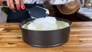 No Bake Cheesecake: Adding whipped cream to a springform pan on a wooden countertop.
