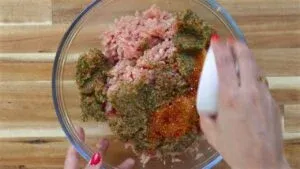 Mixing spices and herbs into ground meat in a glass bowl for a flavorful dish on a wooden surface.