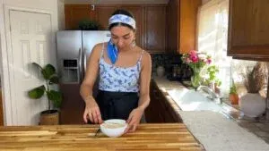 Person in kitchen whisking tahini sauce in bowl, surrounded by plants and flowers.