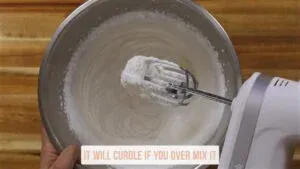 Electric mixer whipping cream in a bowl with a note saying, It will curdle if you over mix it.