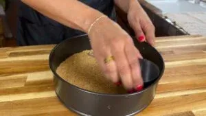 Hands pressing graham cracker crust into a round pan on a wooden countertop.