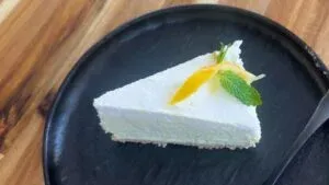 Slice of creamy cheesecake with lemon peel and mint garnish on a black plate, set on a wooden table.