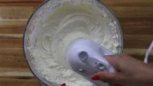 Hand using electric mixer in bowl of creamy batter on wooden surface.