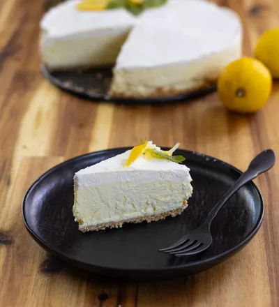 Slice of No-Bake Mascarpone Lemon Cheesecake on a black plate with a fork, garnished with lemon zest and mint, wooden table background.