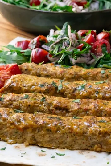 Grilled kebabs on a platter with fresh salad and roasted tomatoes, garnished with herbs. Perfect for a Mediterranean feast.