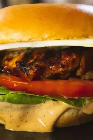Close-up of a grilled chicken sandwich with lettuce, tomato, cheese, and sauce on a toasted bun.