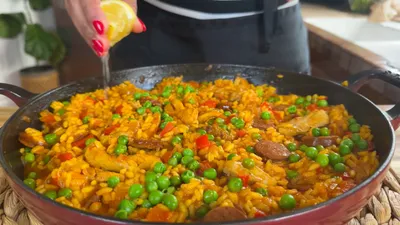 Person squeezing lemon over colorful paella with chicken, sausage, peas, and red peppers in a large pan.