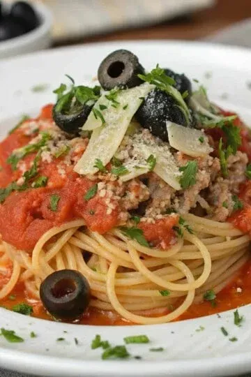 Spaghetti with tuna, tomato sauce, olives, and parmesan, garnished with parsley on a white plate.