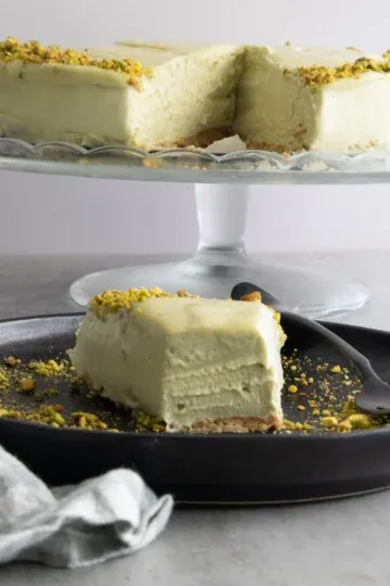 Slice of no bake mascarpone pistachio cheesecake on black plate, garnished with crushed pistachios, on a cake stand.
