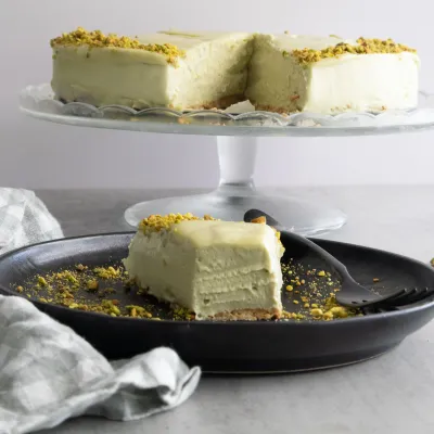 Slice of no bake mascarpone pistachio cheesecake on black plate, garnished with crushed pistachios, on a cake stand.