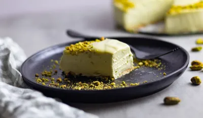 Creamy pistachio cheesecake slice on a black plate, with crushed pistachios on top and around.