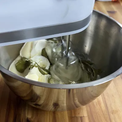 Electric mixer blending cream cheese and pickles in a metal bowl on a wooden countertop.
