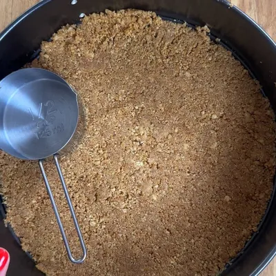 Crushed graham cracker crust in a round pan with a measuring cup for a perfect cheesecake base.