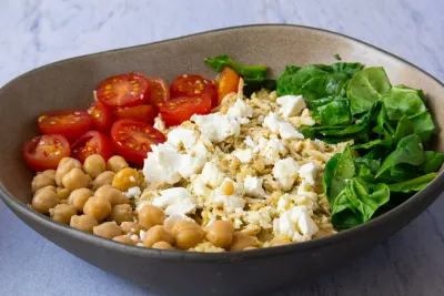 Bowl with chickpeas, cherry tomatoes, feta, spinach, and grains on a stone surface.