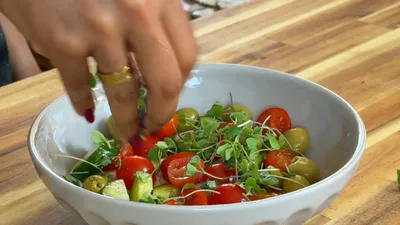 Hand preparing fresh cherry tomato salad with microgreens in a white bowl on wooden table.