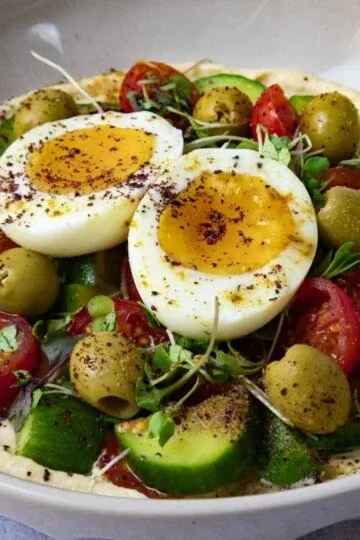 mediterranean hummus bowl with boiled eggs, olives, cherry tomatoes, cucumber, and sprouts on a white plate.