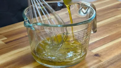 Whisk mixing olive oil in a glass measuring cup on a wooden countertop.