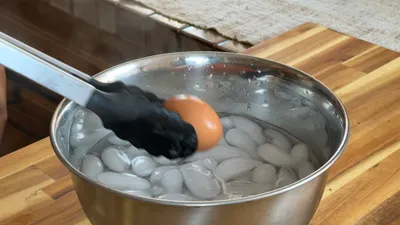 Tongs holding brown egg above ice water bath in metal bowl on wooden surface.