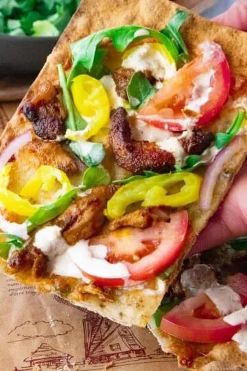 Hand holding a slice of flatbread pizza with tomatoes, peppers, greens, and creamy sauce.