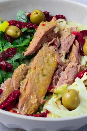 Fresh salad with tuna, arugula, olives, sun-dried tomatoes, yellow peppers, and fennel in a white bowl.