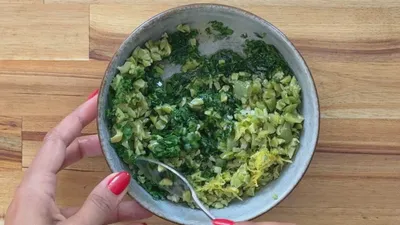 Bowl of chopped green olives, herbs, and lemon zest, stirred with a spoon on a wooden surface.