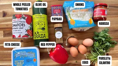 Ingredients for shakshuka: whole peeled tomatoes, olive oil, paprika, onions, harissa, feta cheese, red pepper, eggs, herbs.