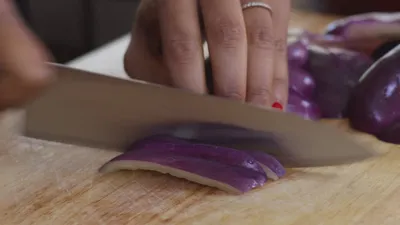 Close-up of a hand slicing a purple bellpepper on a wooden cutting board with a sharp knife.