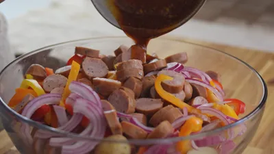 Pouring sauce over sliced sausage, onions, and peppers in a glass bowl.
