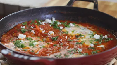 Pan of shakshuka with poached eggs in spicy tomato sauce, garnished with herbs and crumbled feta cheese.